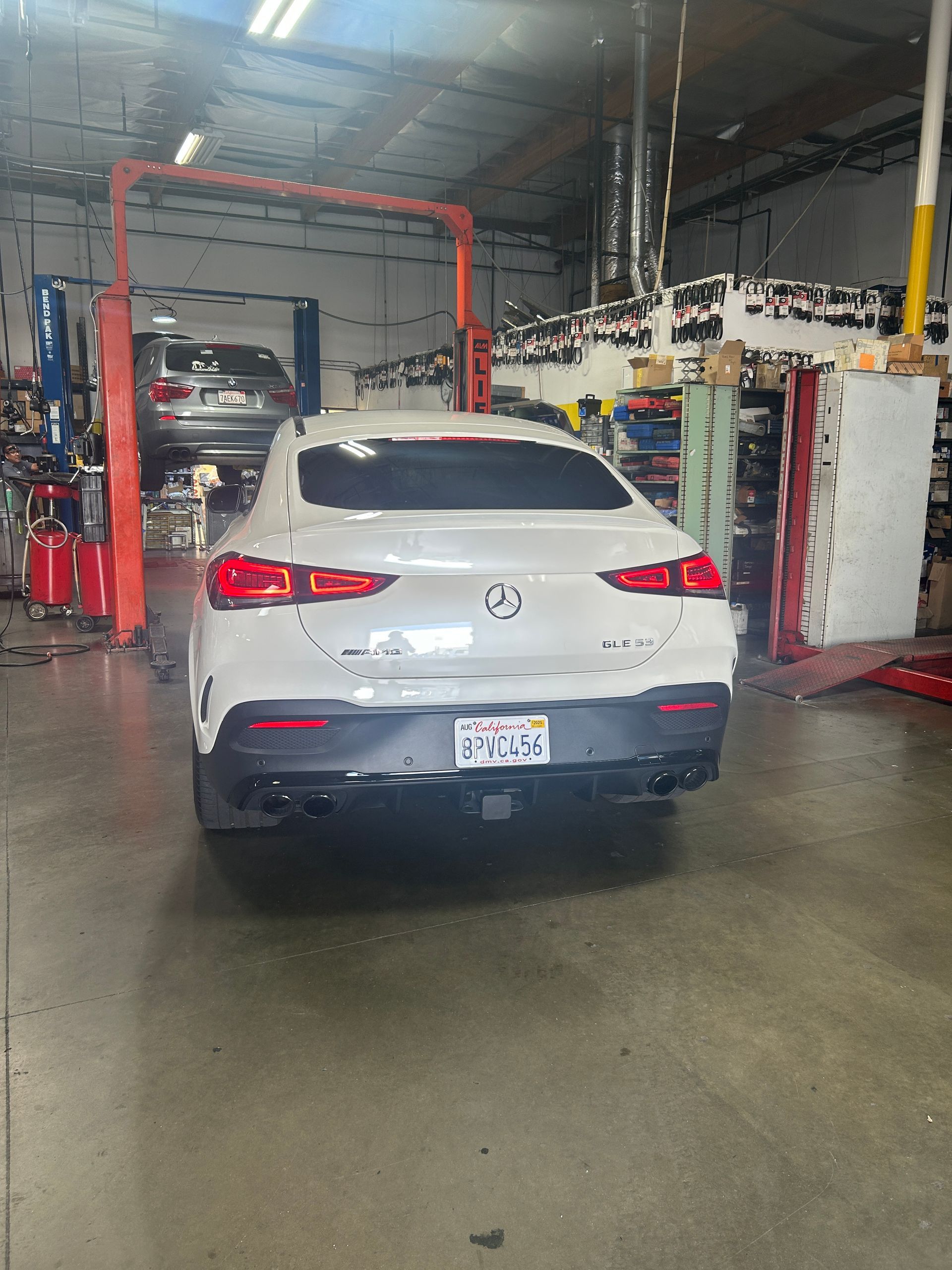 White SUV in an auto repair shop bay, seen from the rear with other vehicles and equipment nearby.