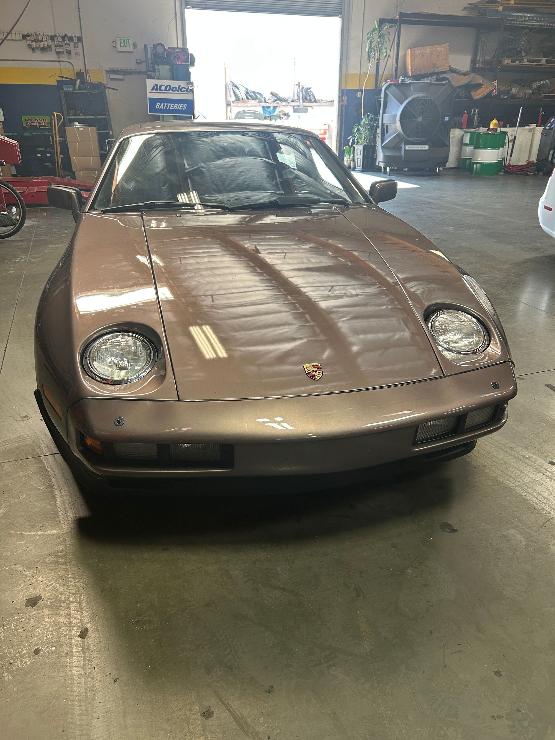 Front view of a tan vintage sports car parked in a garage, with a bright open doorway behind it