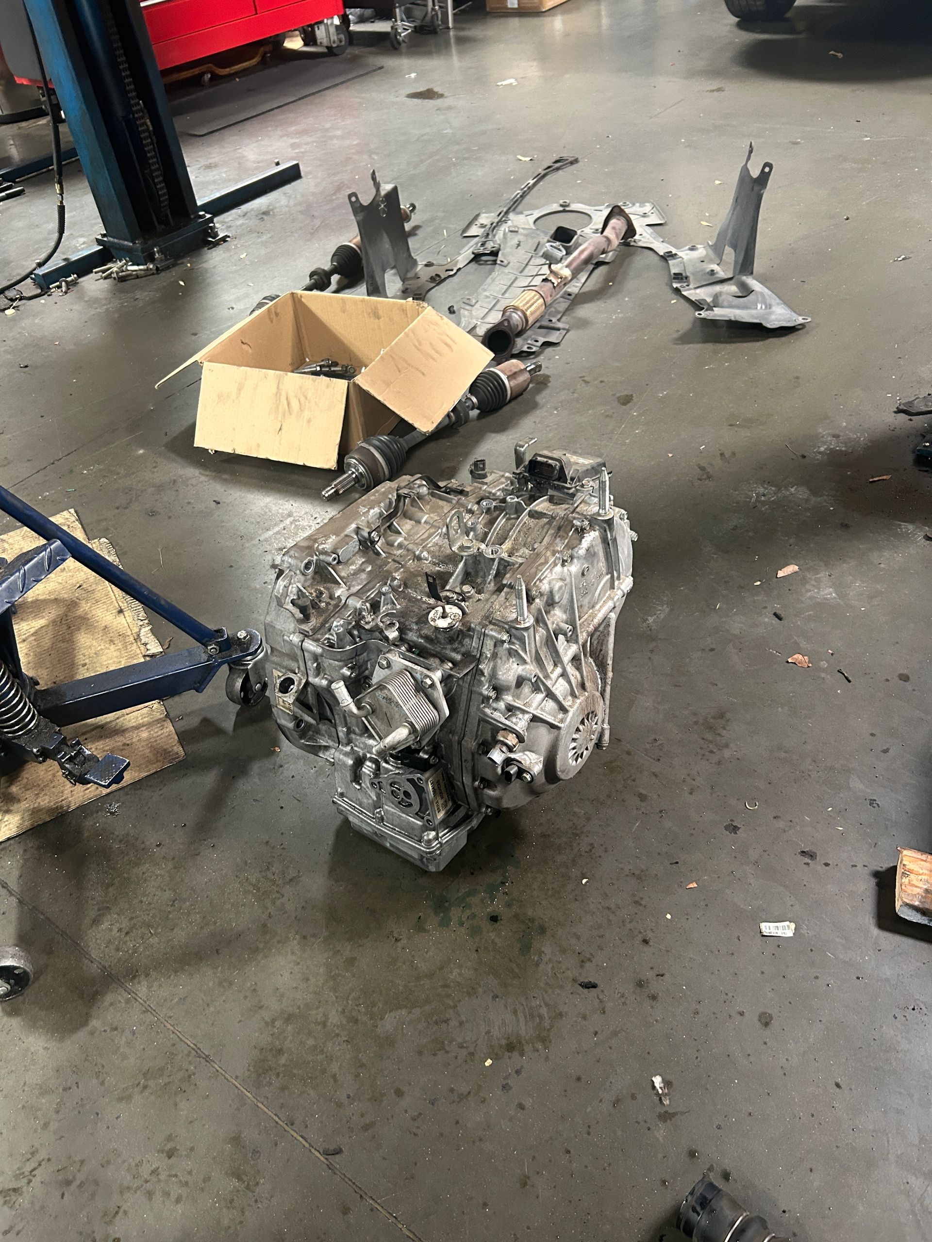 Damaged car transmission on a workshop floor beside a cardboard box and scattered parts