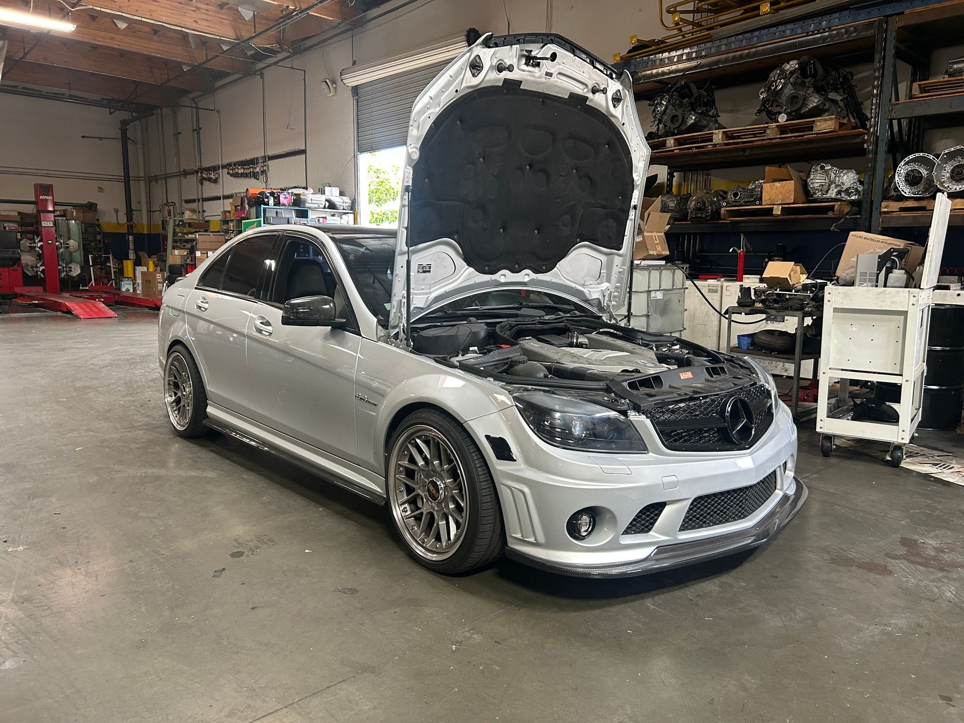 Silver car with hood open in an auto repair shop, showing the engine bay.