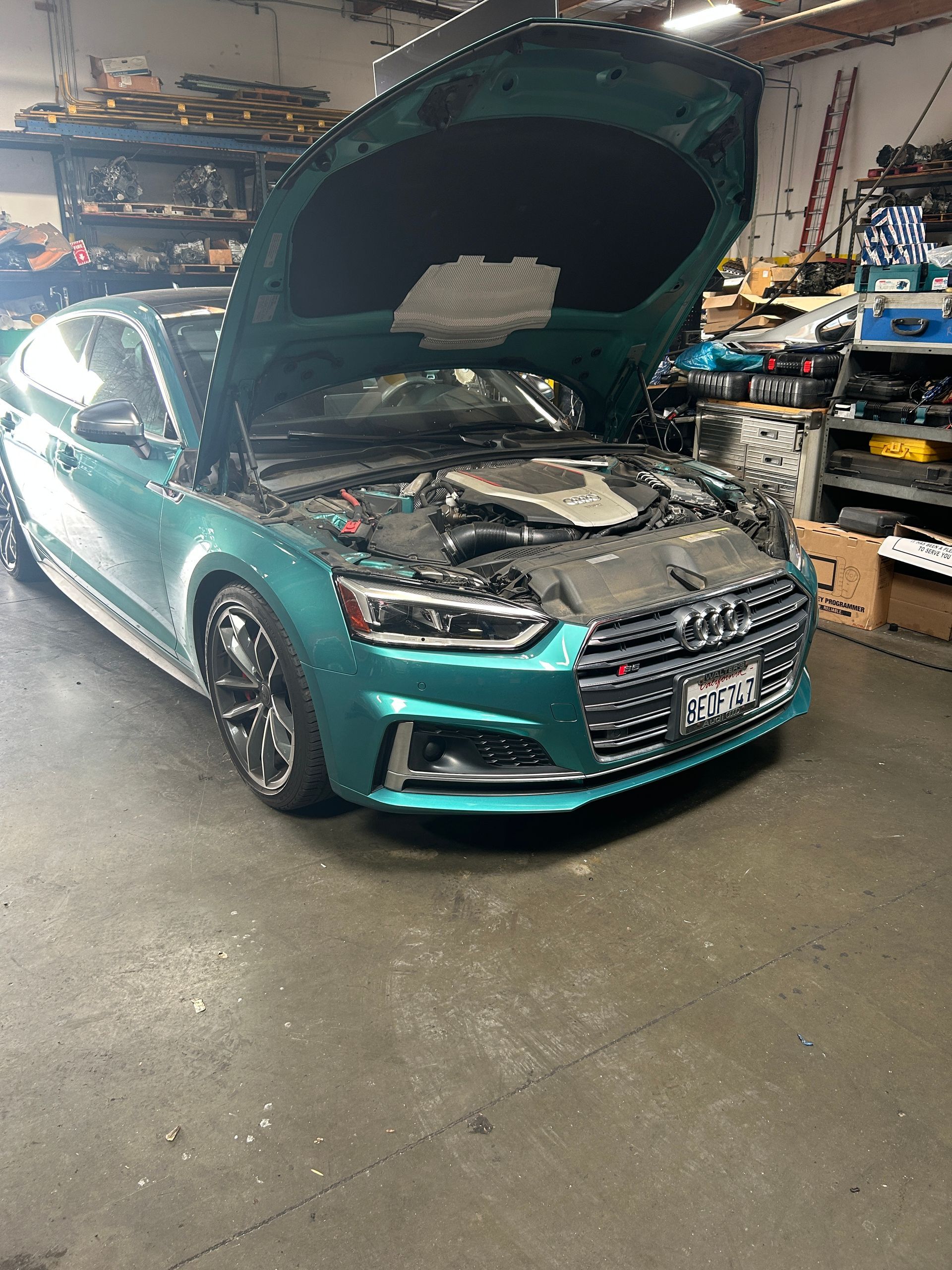 Teal car with hood open in a mechanic’s garage, engine exposed.
