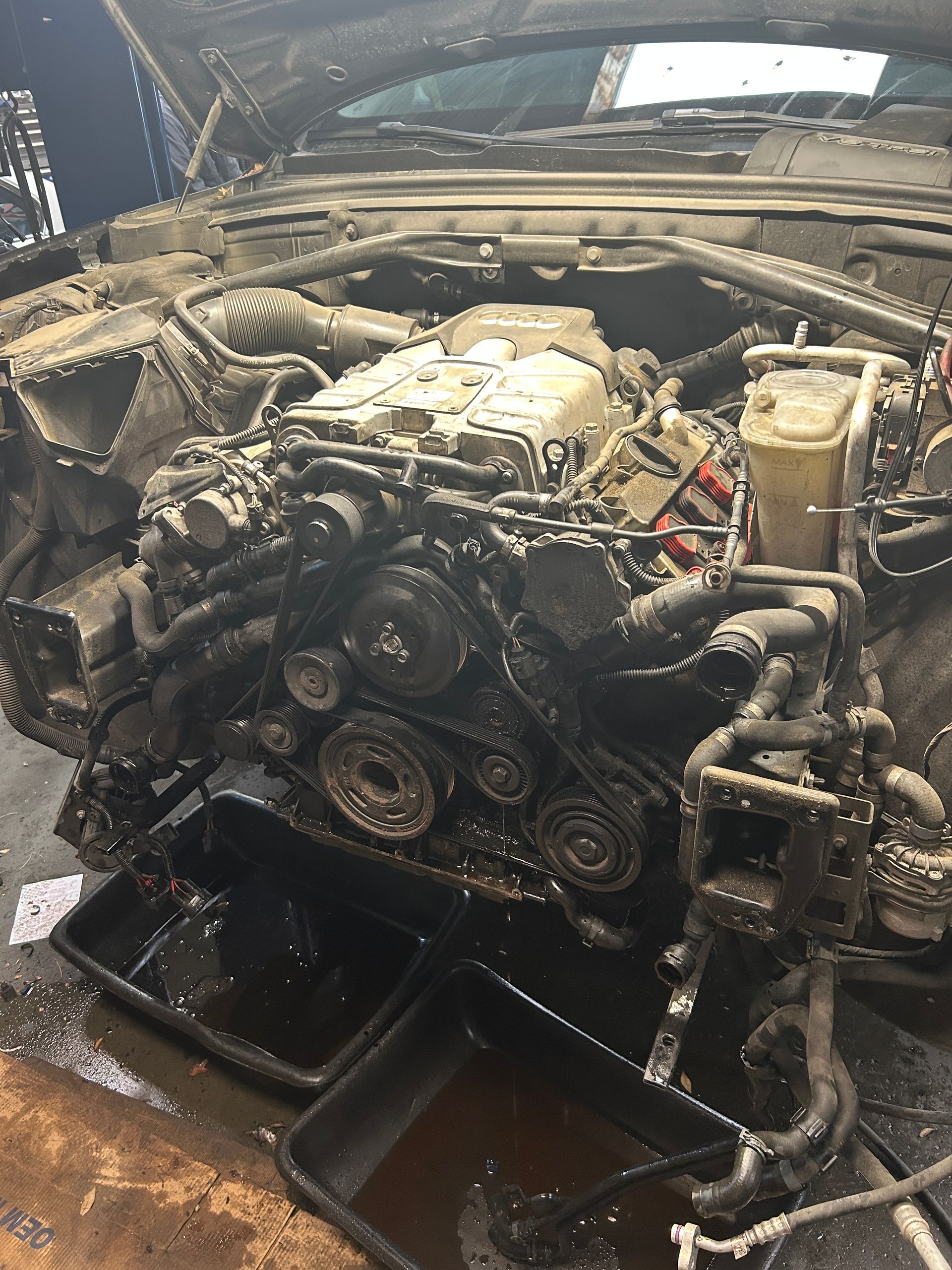 Dirty car engine with exposed belts and hoses, sitting above a drain pan in a repair bay.
