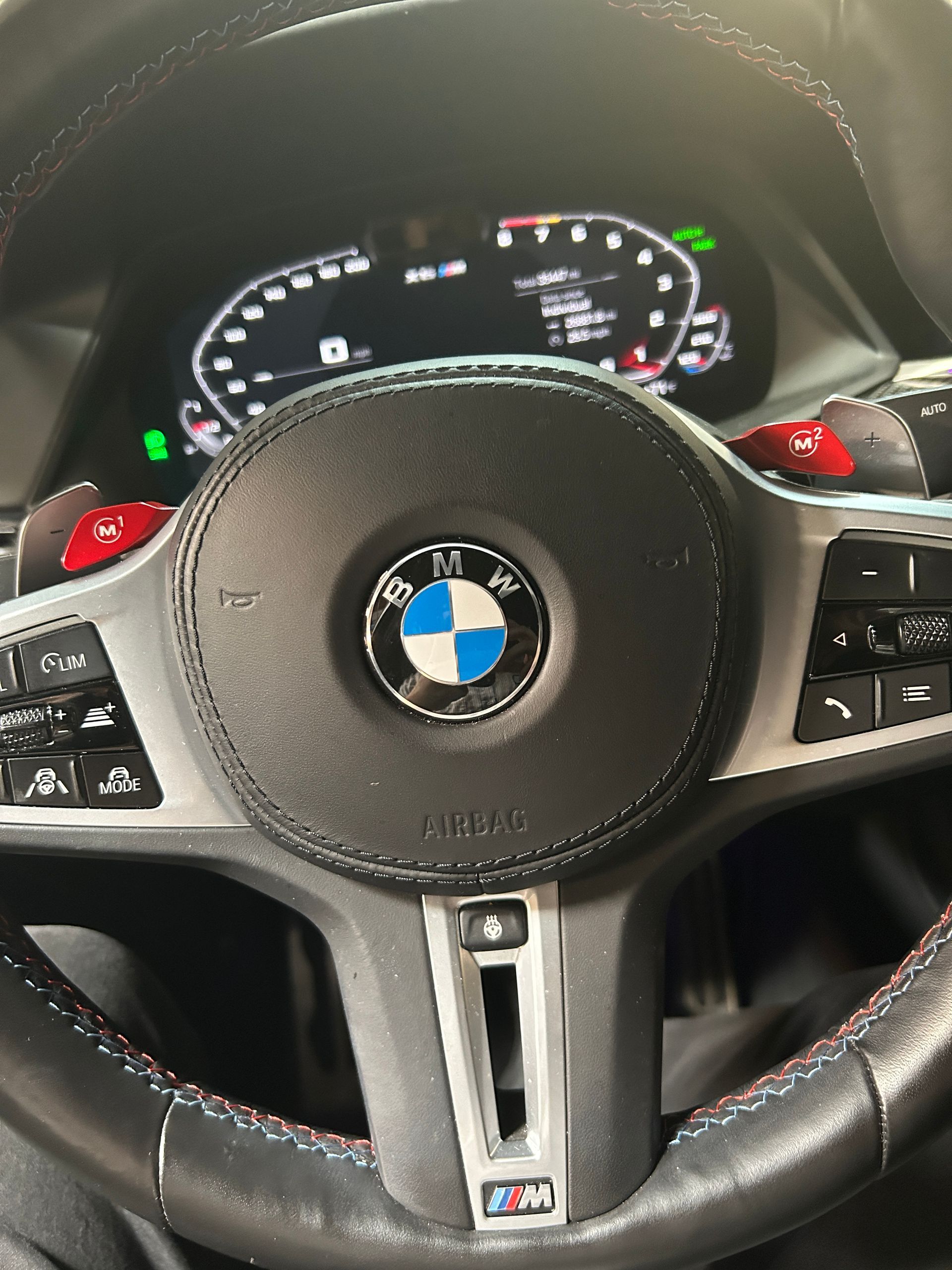 BMW steering wheel and digital dashboard inside a car, with red stitching and illuminated controls.