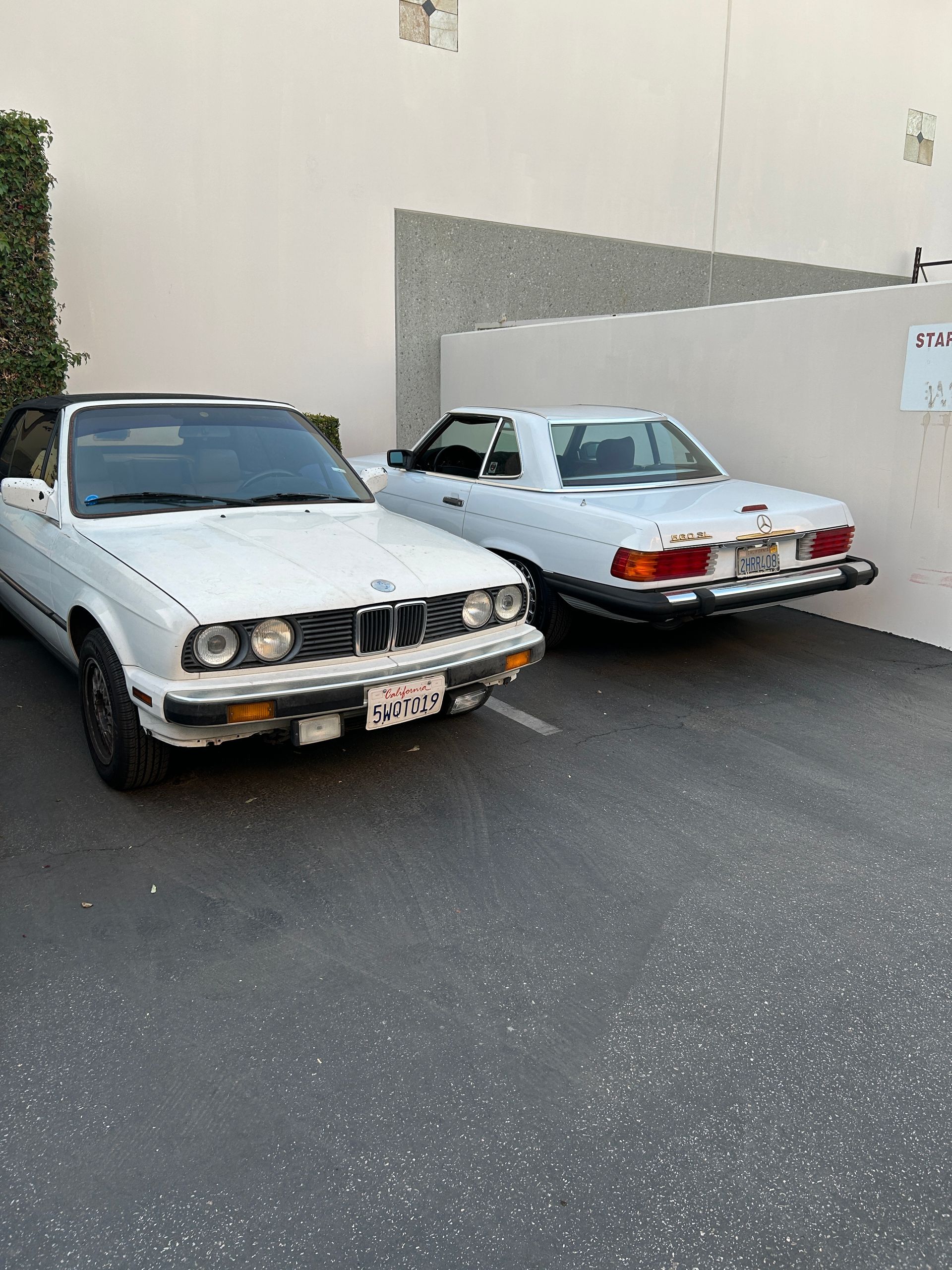 Two white vintage convertibles parked beside a beige wall in a parking lot.
