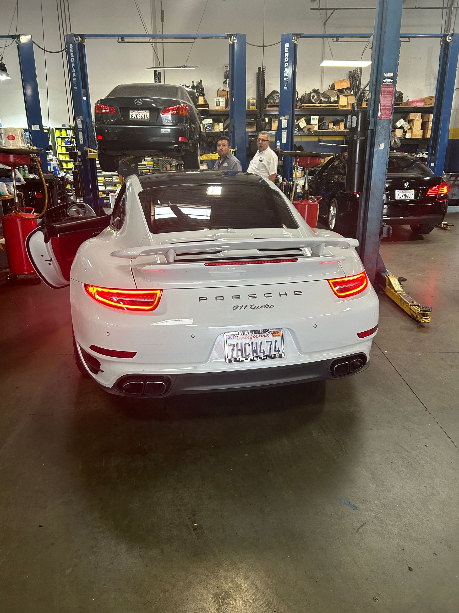 White sports car in an auto repair shop, seen from the rear with brake lights on and another car on a lift nearby