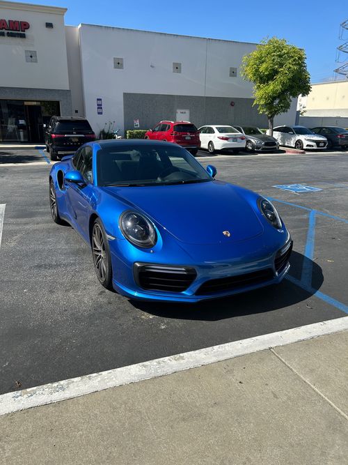 Blue Porsche sports car parked in a sunlit lot outside a white building.