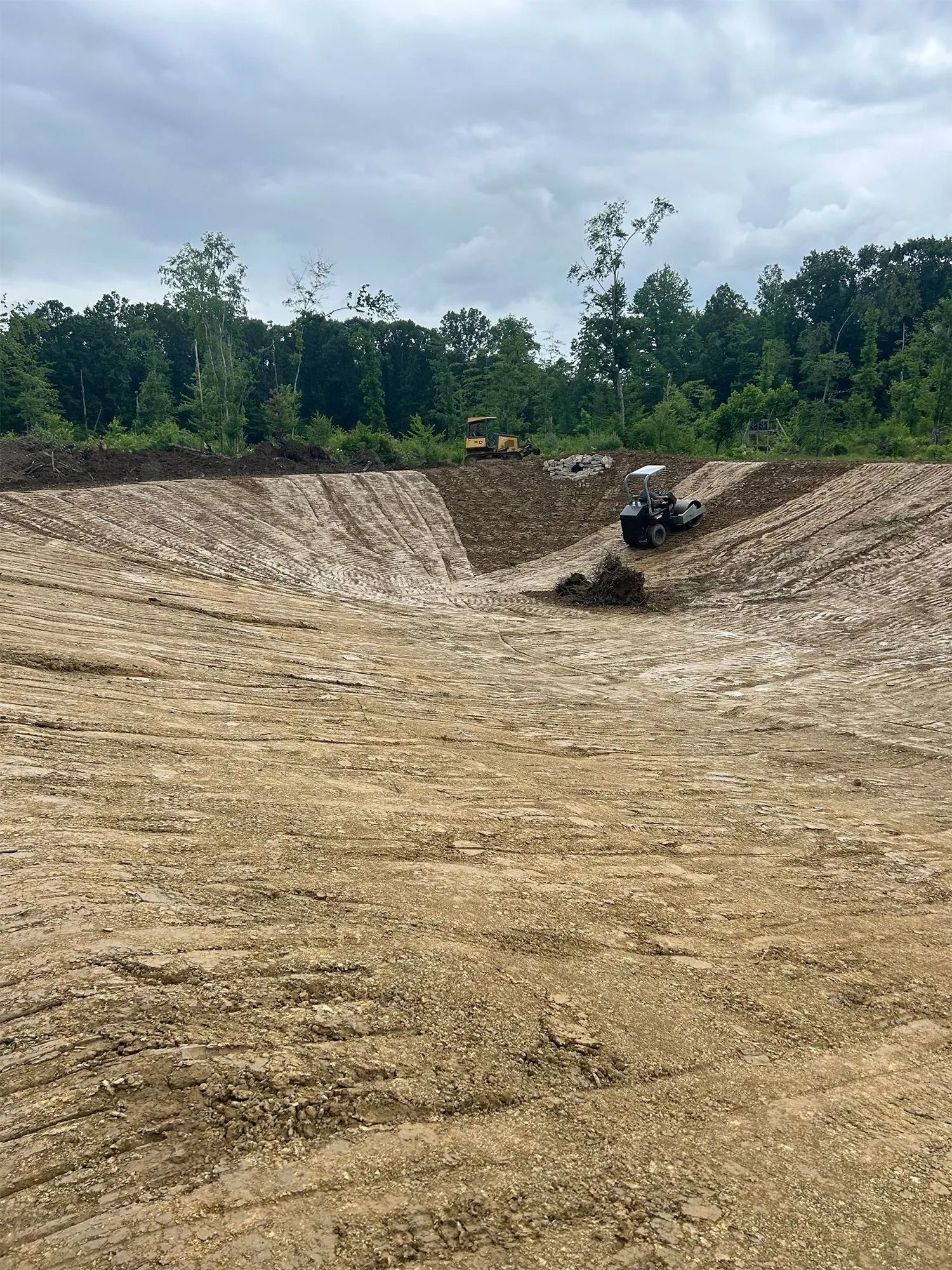Construction site: Excavator and tractor grading dirt for a pond against a tree line. Cloudy sky.