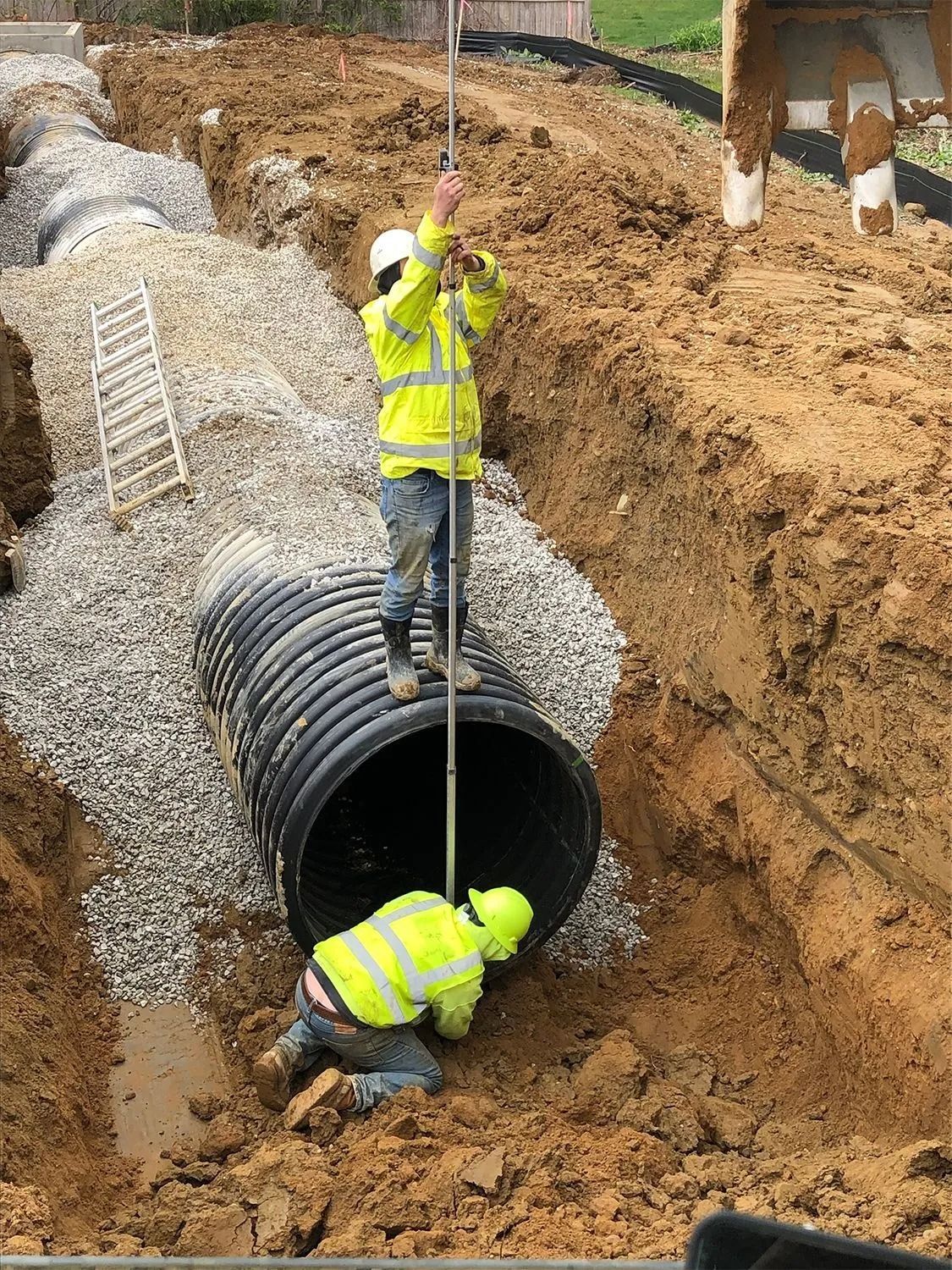 Construction workers inspecting a large drainage pipe in an excavation, one using a measuring rod.