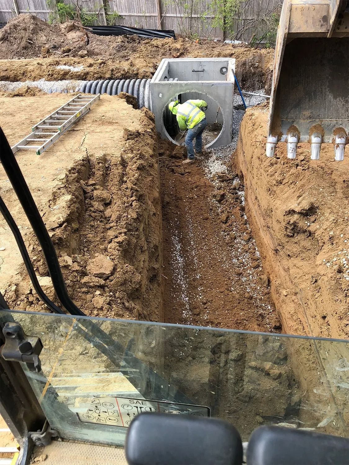 Construction worker in trench near concrete drainage box and excavator.