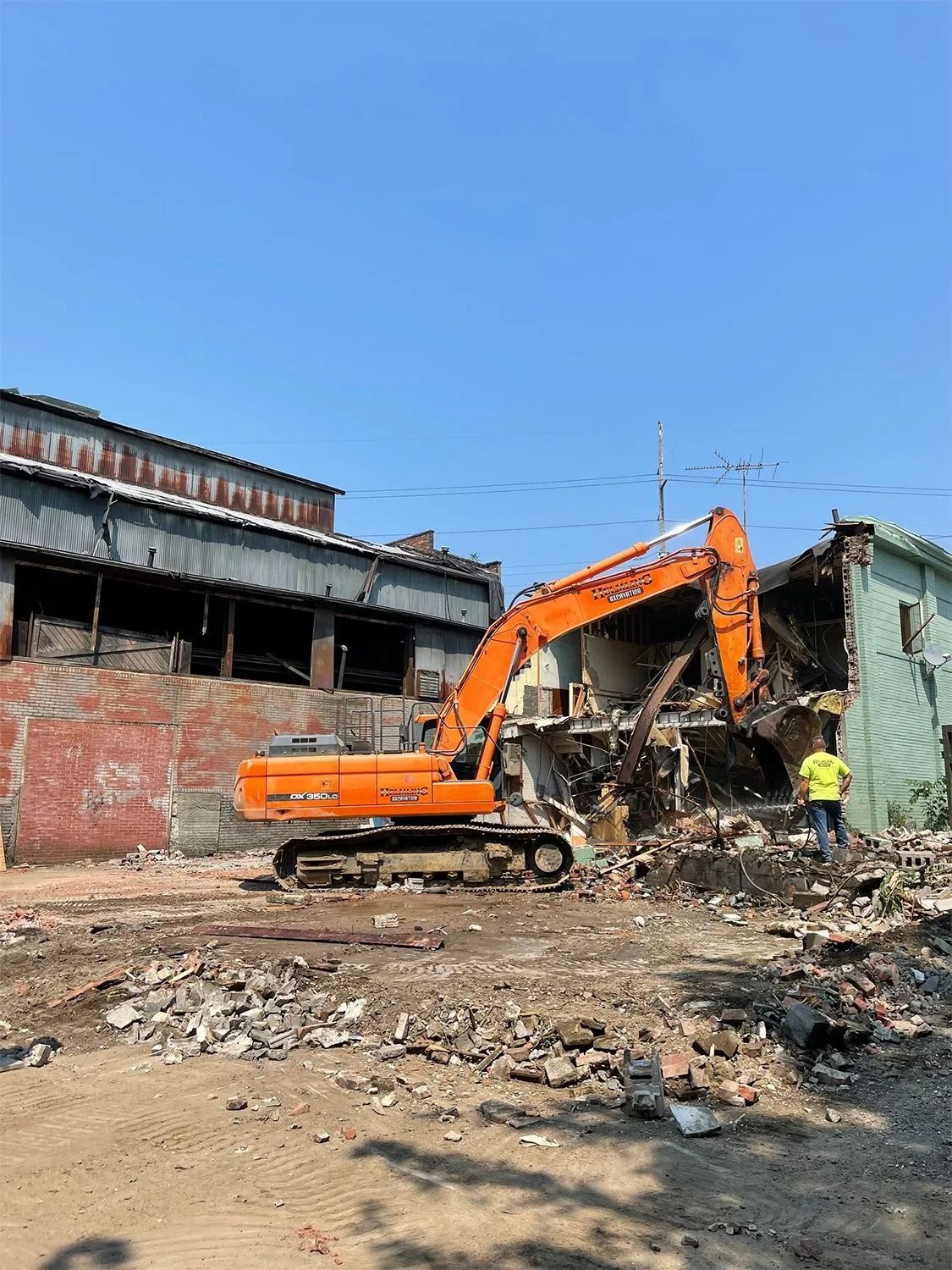 Orange excavator demolishing a building under a blue sky, with a worker in a yellow vest nearby.