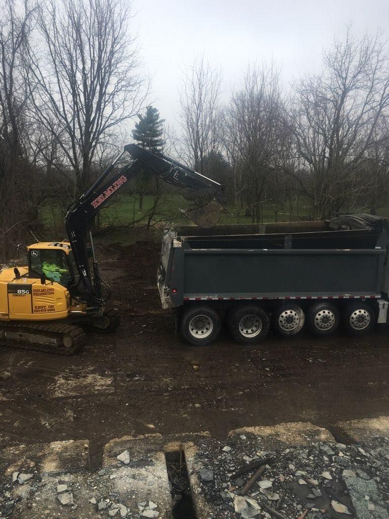 Yellow excavator loading a gray dump truck with dirt on a construction site, trees in the background.