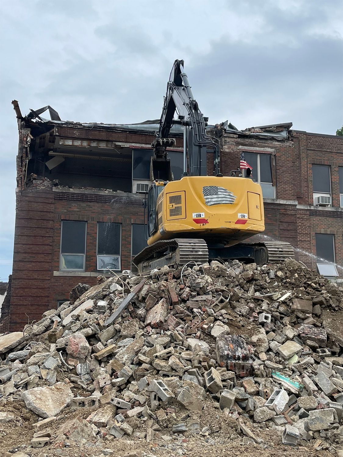Excavator demolishing brick building, perched atop debris pile on a cloudy day.