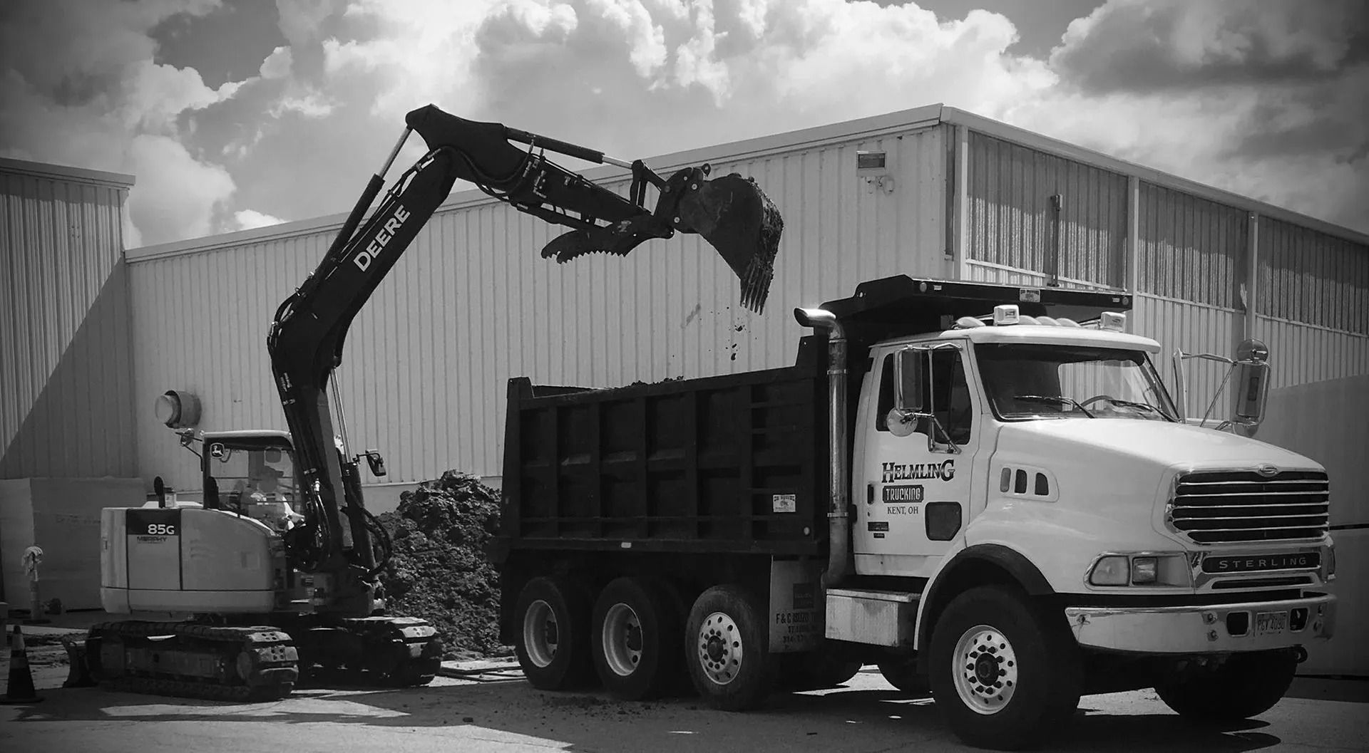 Excavator loading dirt into dump truck in front of a white industrial building.
