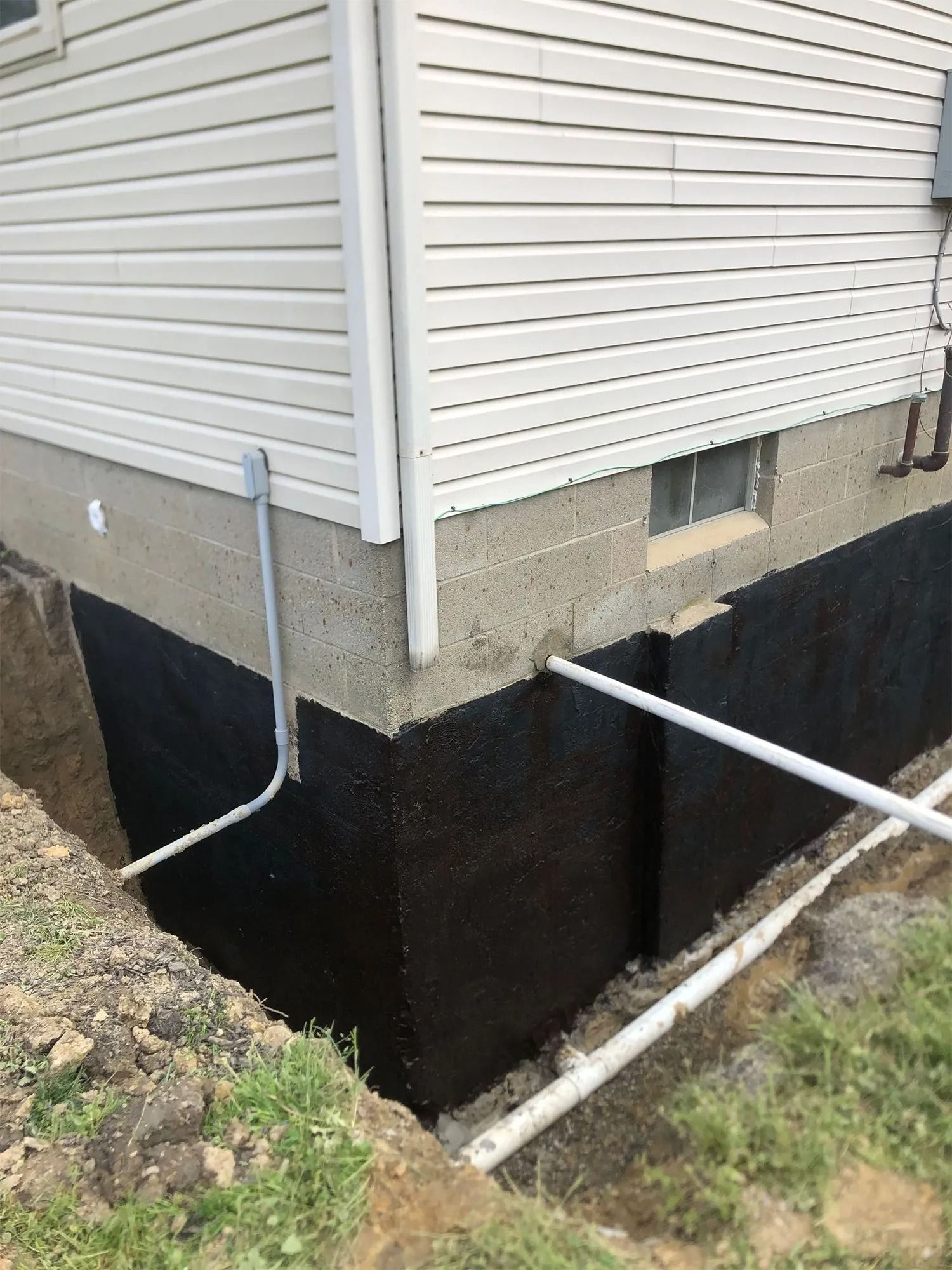 Black waterproofing applied to a foundation corner; white siding above; exposed soil at the base; drain pipes.