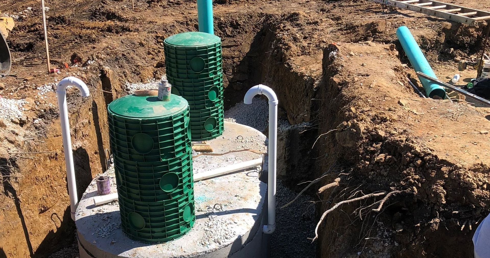 Two green septic tank risers in an excavated pit, with white PVC pipes nearby.