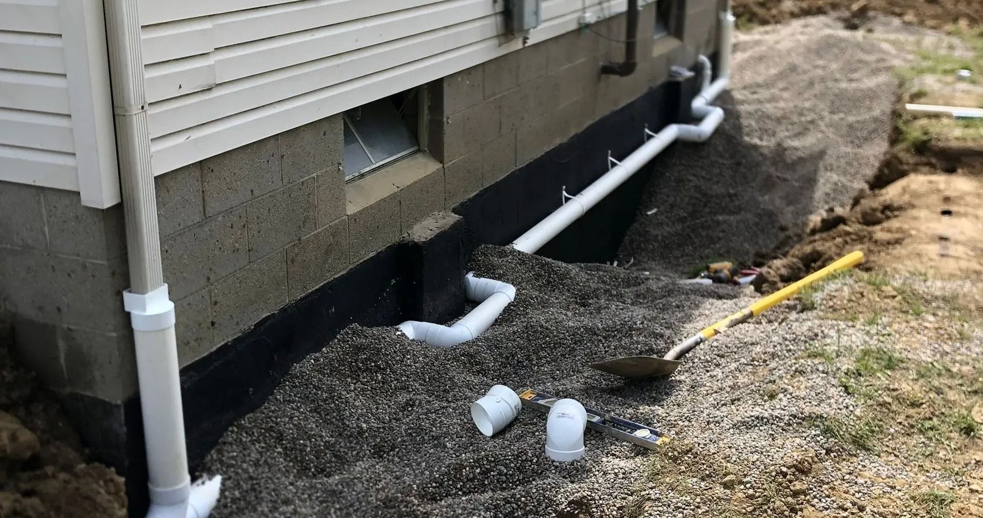 Foundation work in progress; exterior house wall with exposed concrete and gravel, white drainage pipes.