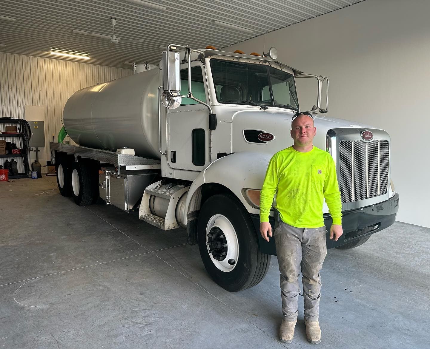 A person in a bright yellow long-sleeved shirt stands in a garage next to a large white tanker truck.