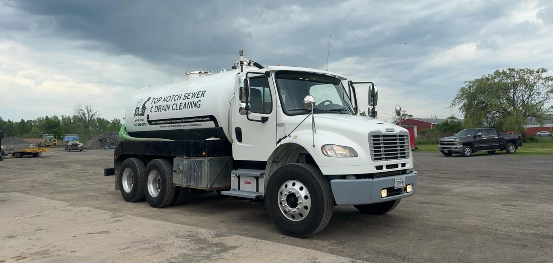 A white industrial vacuum truck with a tank sits parked on a gravel lot under a cloudy sky.