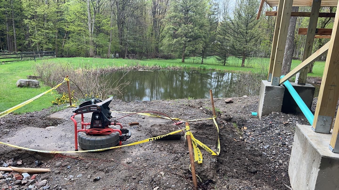 A small pond behind a building construction site, featuring a red machine on a tire and yellow safety tape in the yard.