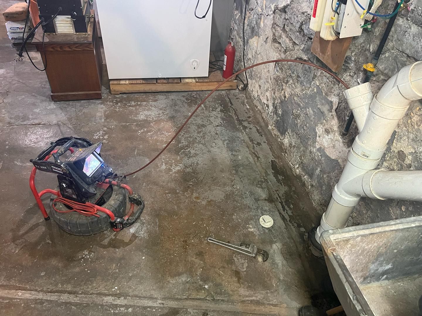 A red drain snake machine on a concrete basement floor, with its cable extending toward a white PVC pipe assembly.