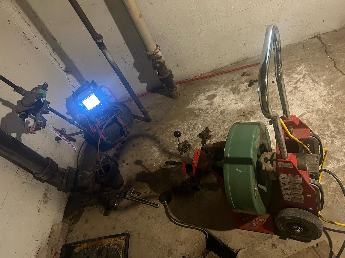 A sewer inspection camera and a red drain cleaning machine are set up on a concrete floor in a basement.
