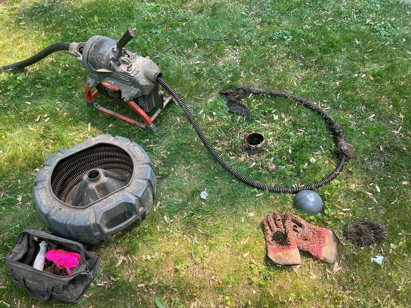 A plumbing snake machine, a plastic drain cover, and work gloves are arranged on a green lawn.