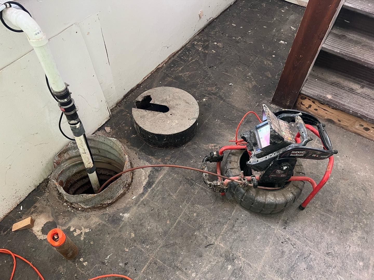 A plumbing drain inspection camera on wheels sits next to an open, concrete-lined sump pit in a dark, industrial space.