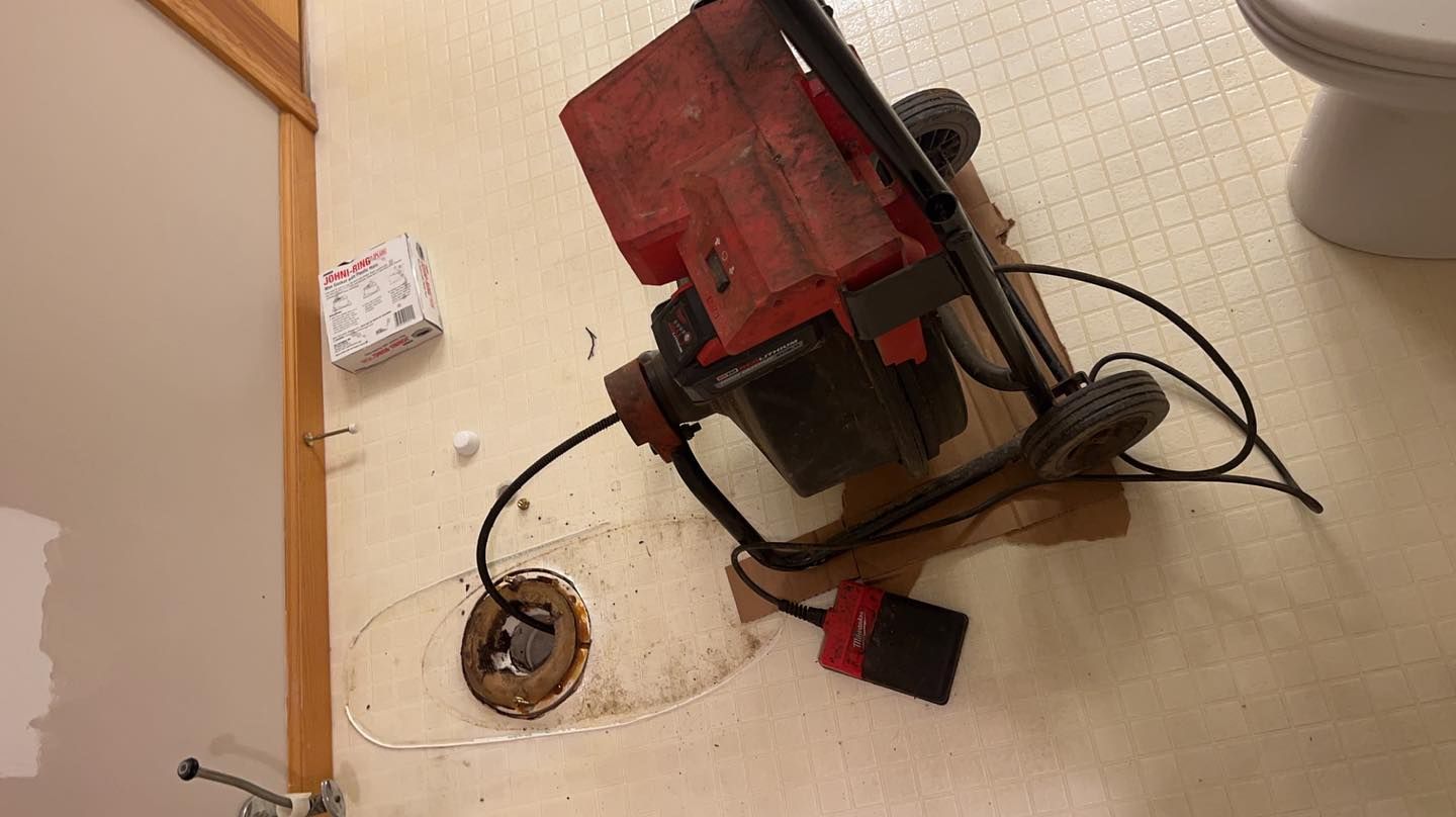 A plumbing snake machine is positioned over a bathroom floor drain, with its cable extending into the pipe.