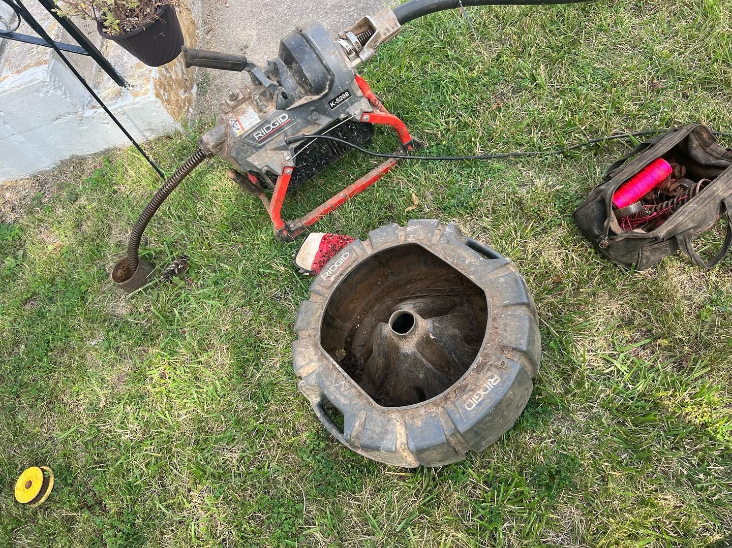 A drain cleaning machine with a red frame sits on grass next to a discarded, circular metal pipe access cover and tool bag.