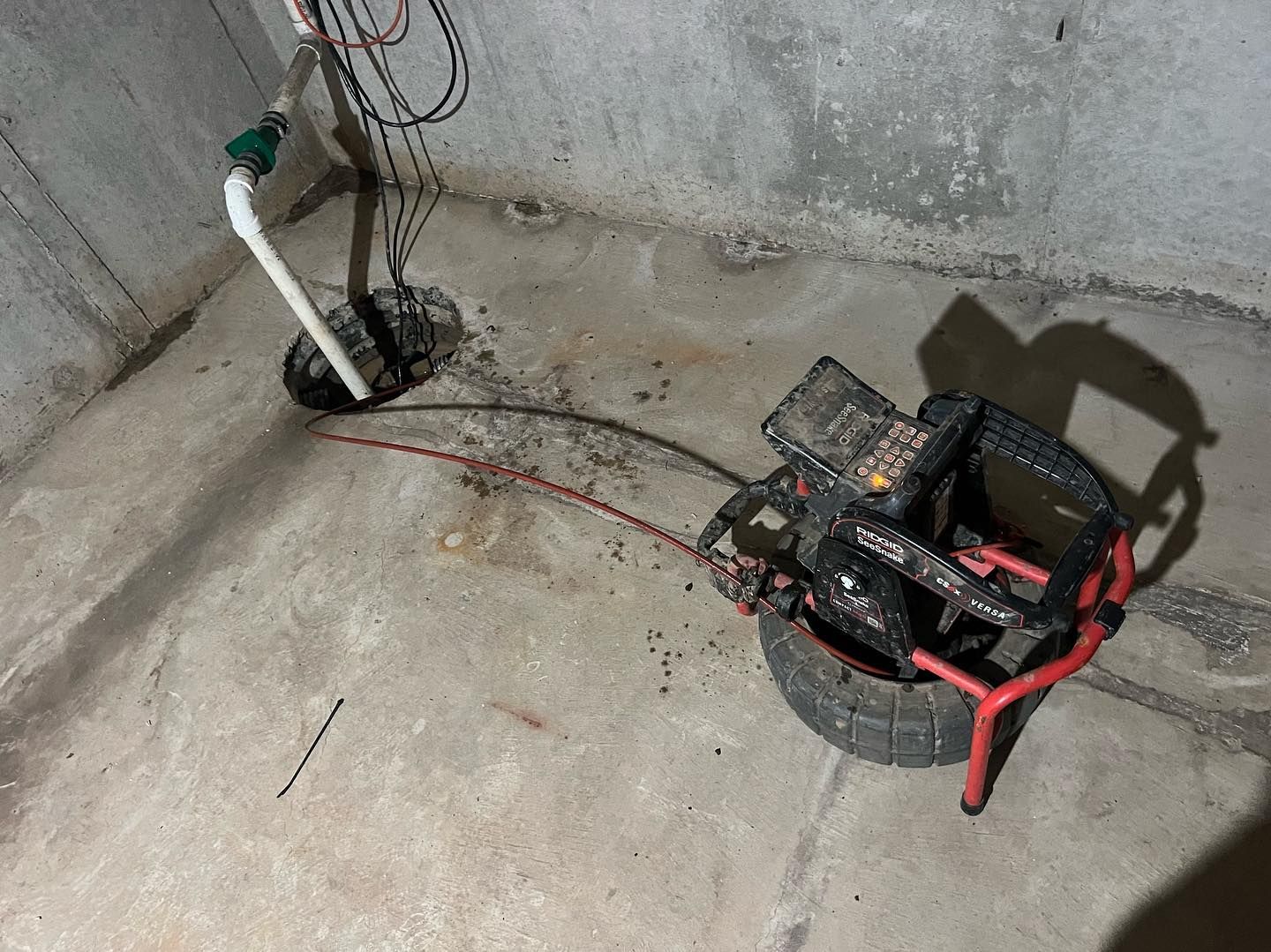 A drain cleaning machine sits on a basement floor, with its cable running into a sump pump pit.