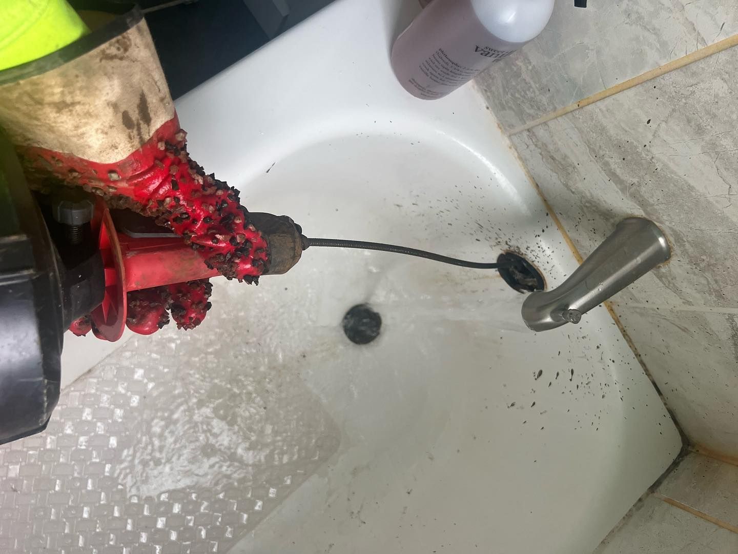 A person wearing work gloves operates a drain snake tool into a bathtub drain, with water running from the faucet.