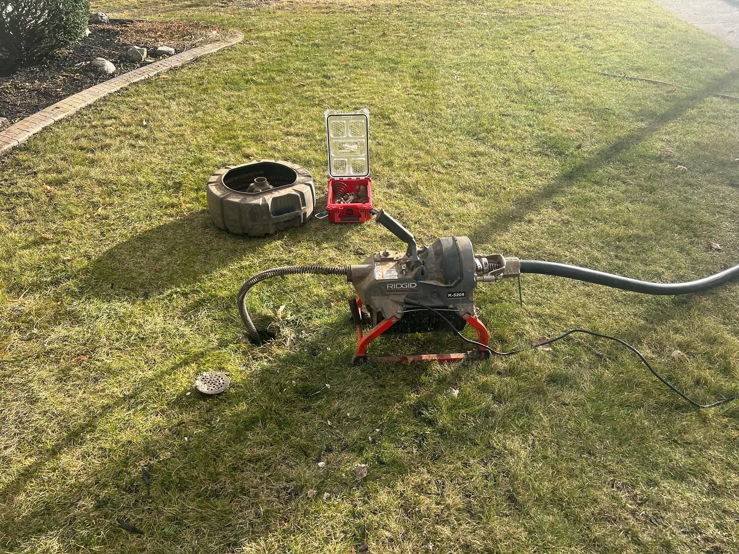 A drain cleaning machine with a power cable is set up on a grassy lawn next to a small red toolbox and an open drain.