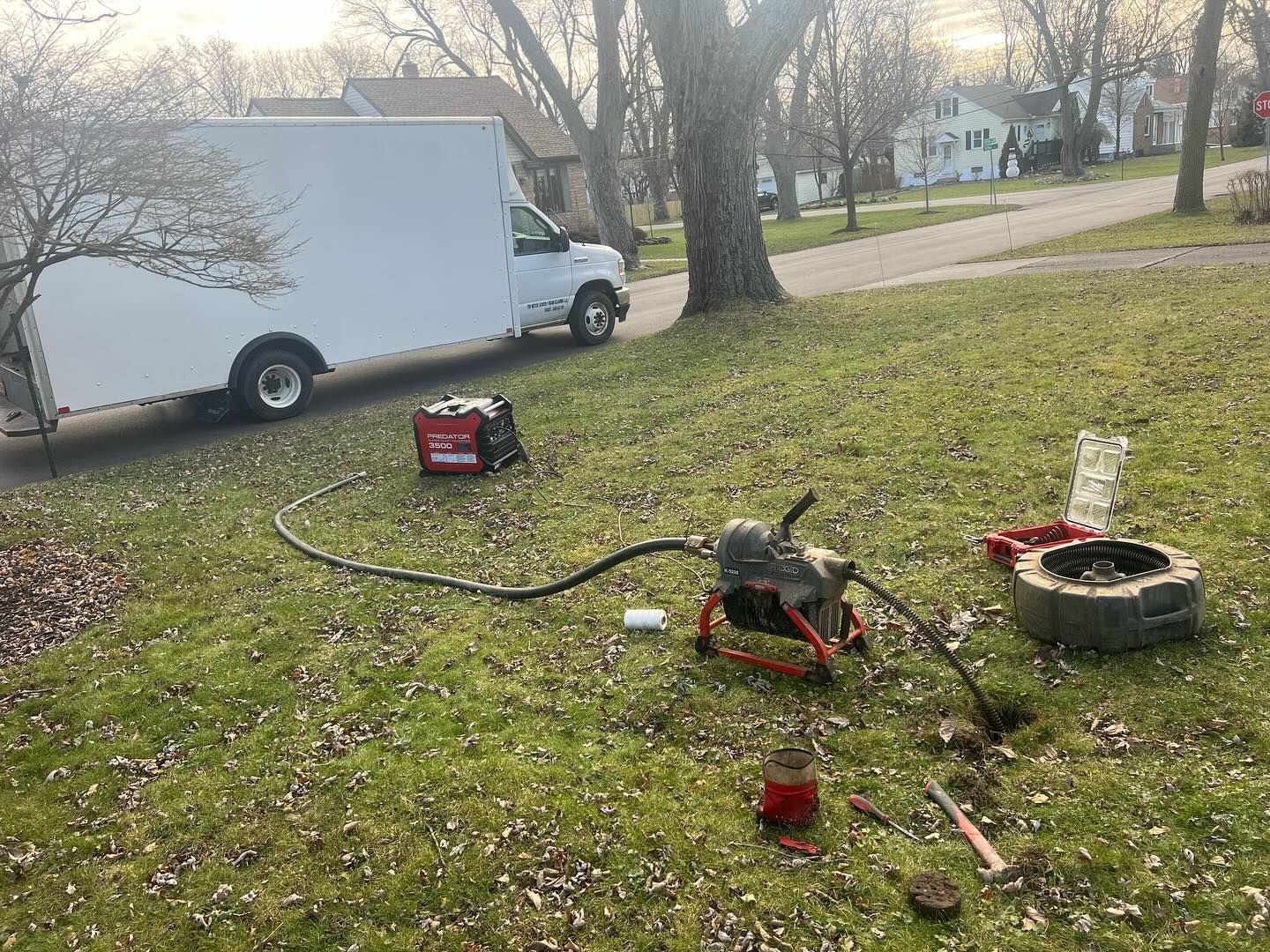 A white utility truck parked on a residential lawn, with drain cleaning equipment connected to an open access point nearby.