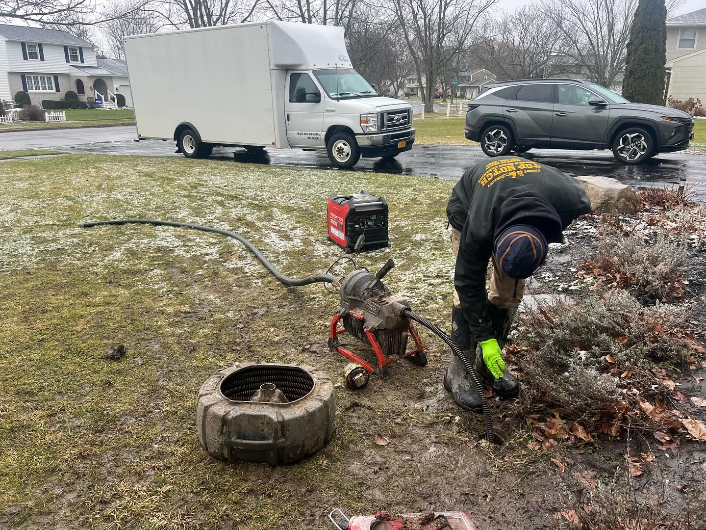 A worker in a dark jacket operates a sewer cleaning machine on a snowy residential lawn, near a truck and parked car.