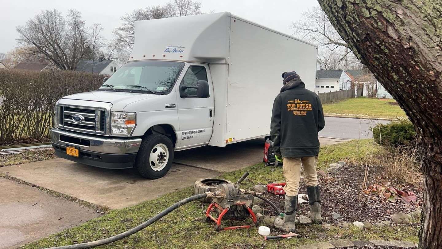 A person in work gear stands near a parked white box truck, attending to equipment on the grass in a residential yard.