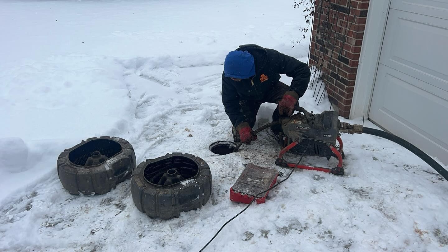 A technician in a blue hat and dark jacket uses specialized plumbing equipment to clear a drain pipe in snowy conditions.