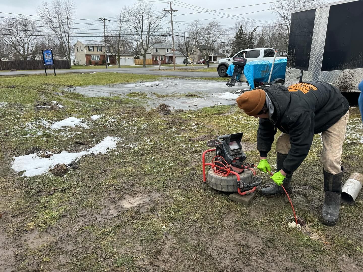 A worker in a black jacket and brown beanie uses a drain cleaning machine on a muddy residential lot.