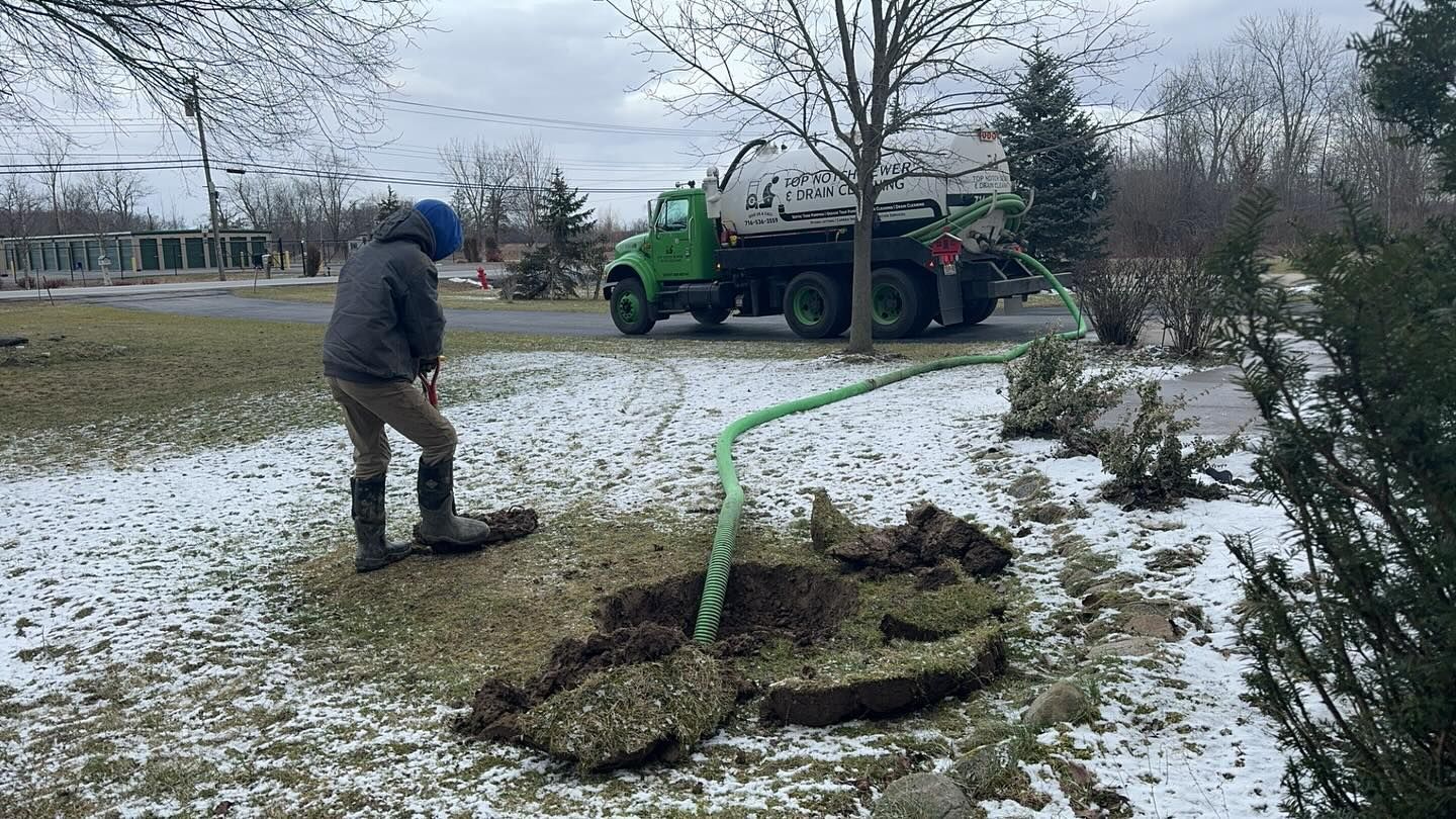 A worker in a gray jacket uses a long green hose connected to a service truck to pump a hole in a snowy, grassy lawn.