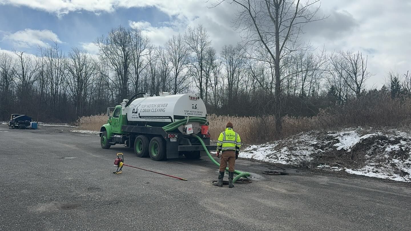 A worker in a yellow high-visibility jacket operates a hose connected to a green tank truck on a gravel lot near trees.