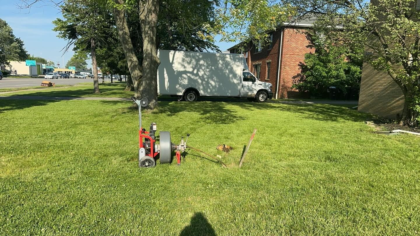 A red industrial drain cleaning machine sits on a grassy lawn next to a white box truck parked by a brick building.