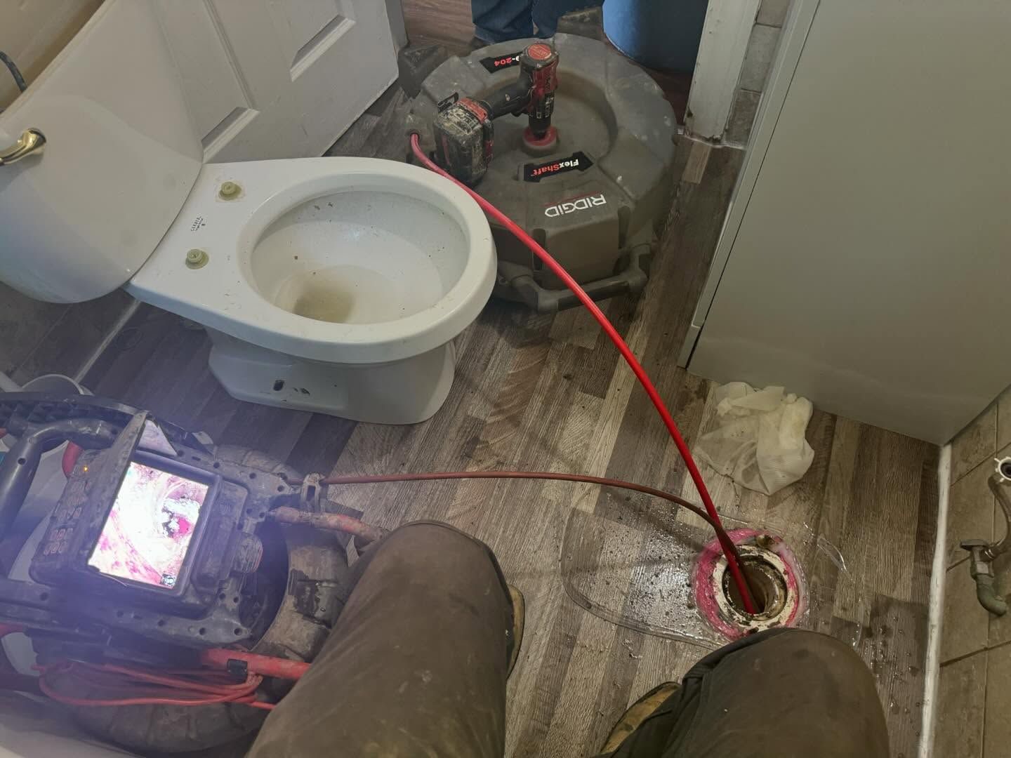 A plumber inspects a bathroom drain using a specialized camera system with a monitor while the toilet is removed.
