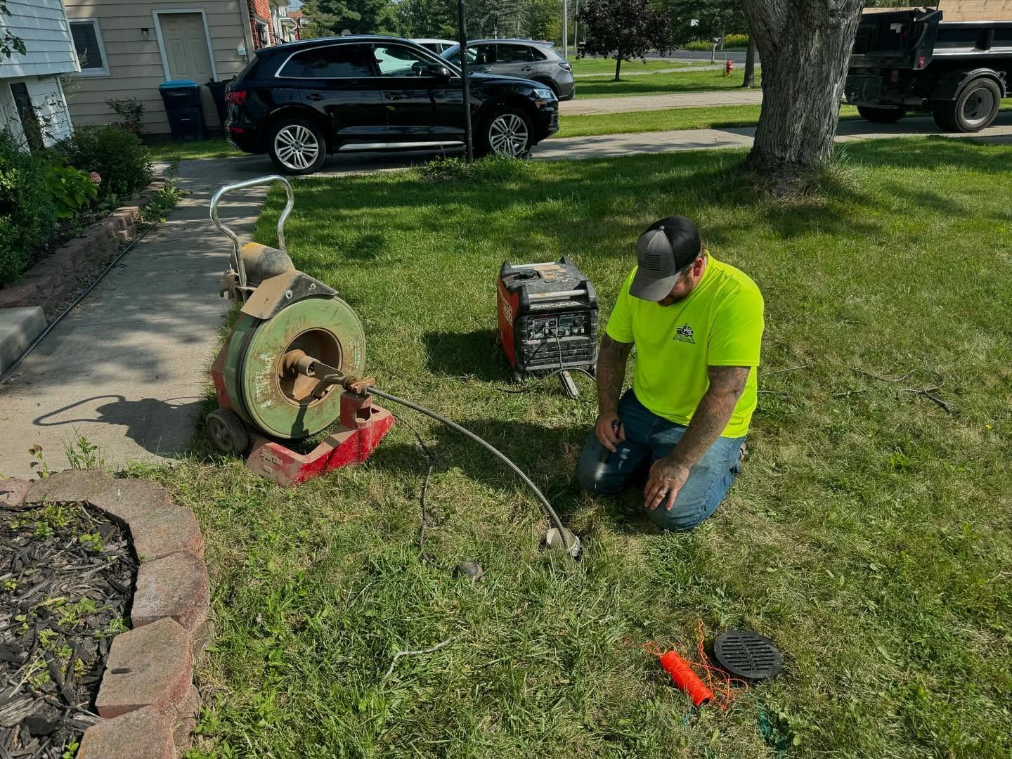 A worker in a neon yellow shirt kneels on a lawn, operating a sewer snake machine to clear an underground pipe.