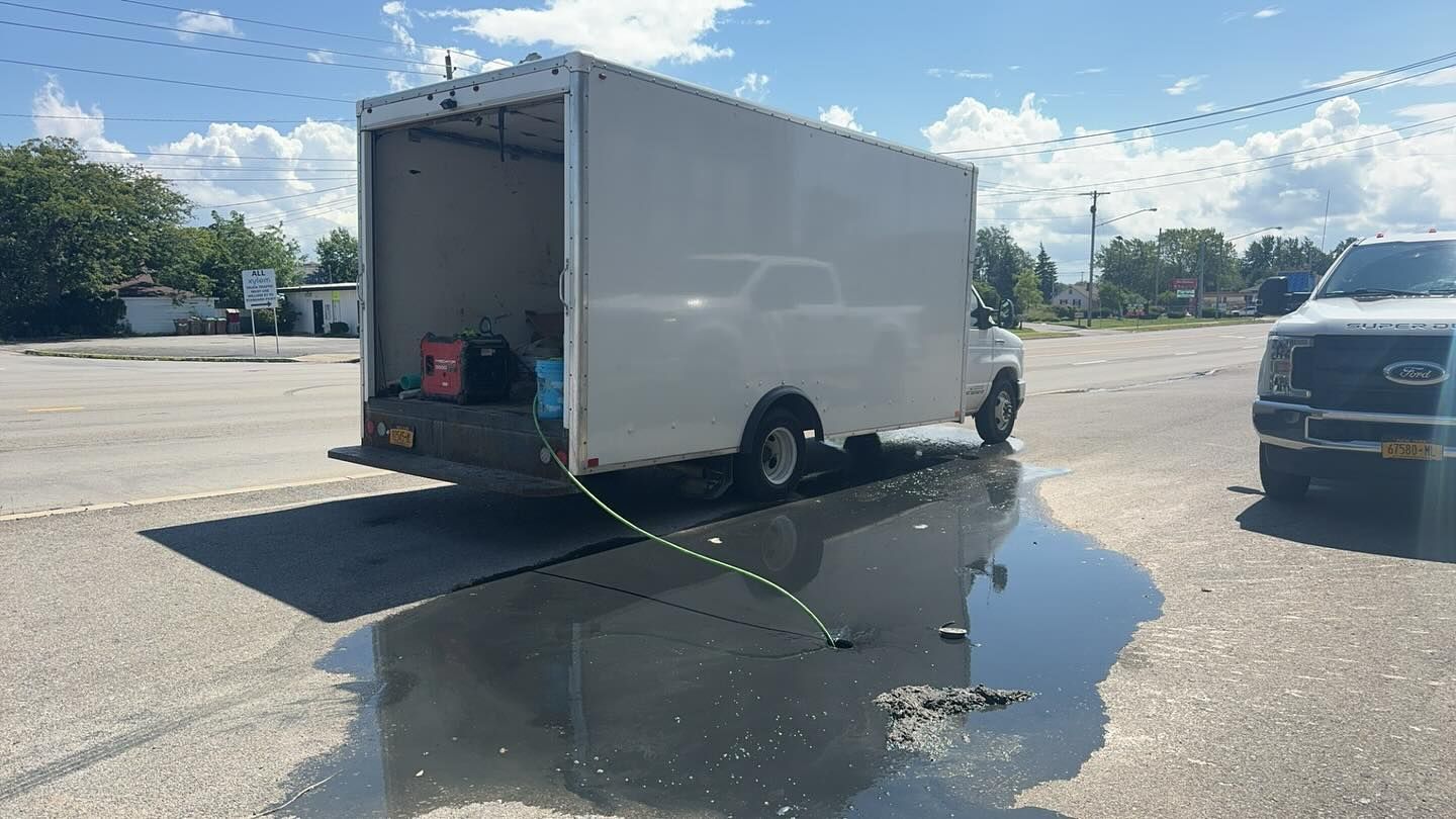 A white box truck parked on a paved lot with a puddle beneath the open rear door and a white truck visible on the right.