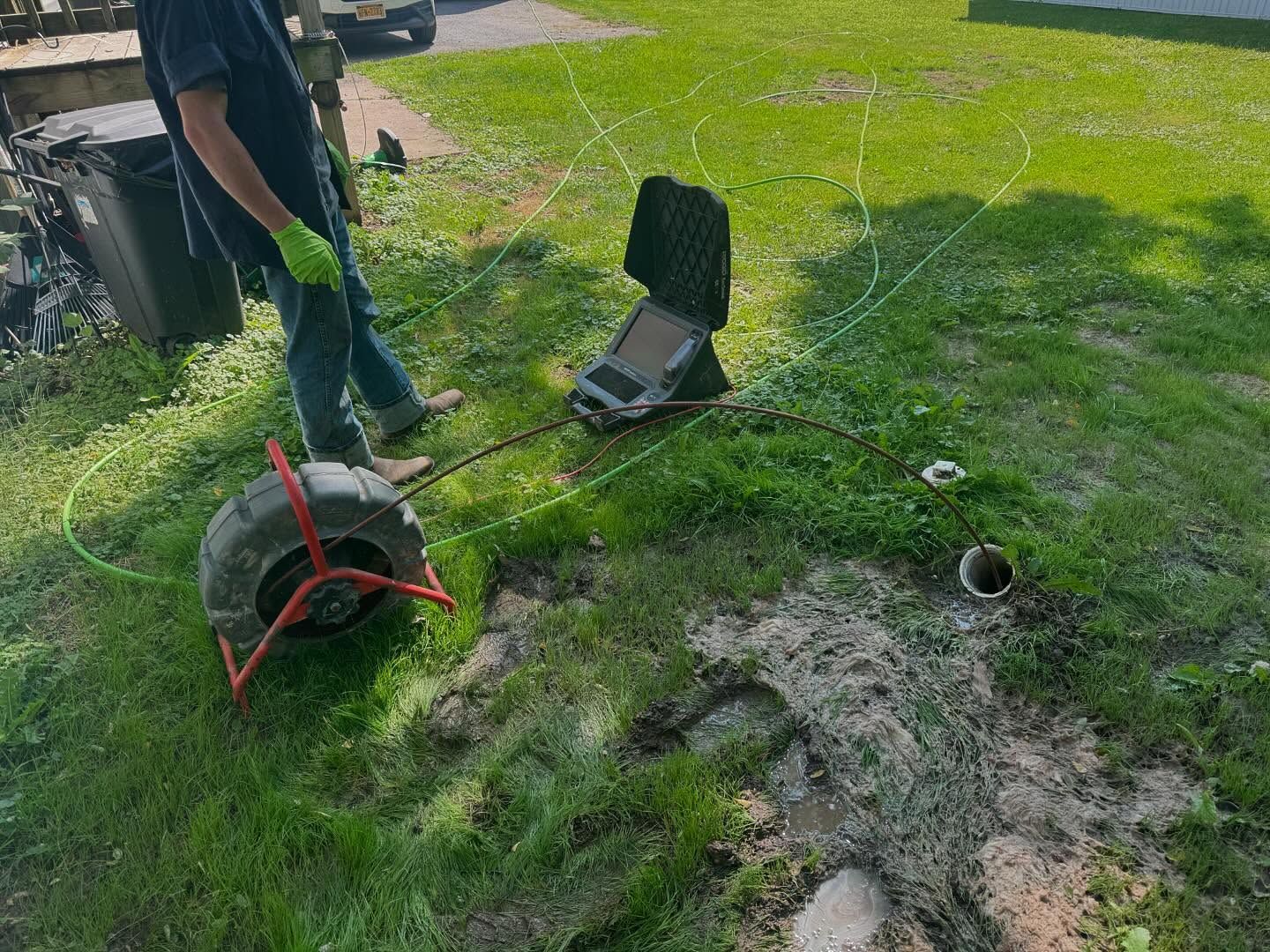A technician operates a sewer camera inspection system on a lawn, with a monitor and cable reel connected to a pipe.