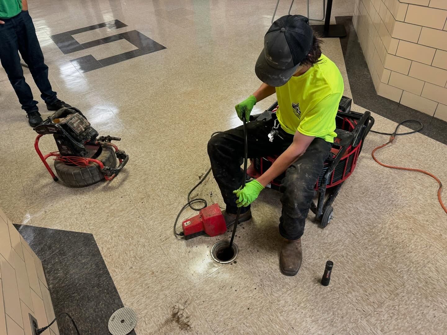 A worker in a bright yellow shirt uses specialized equipment to inspect a drain in a hallway floor.