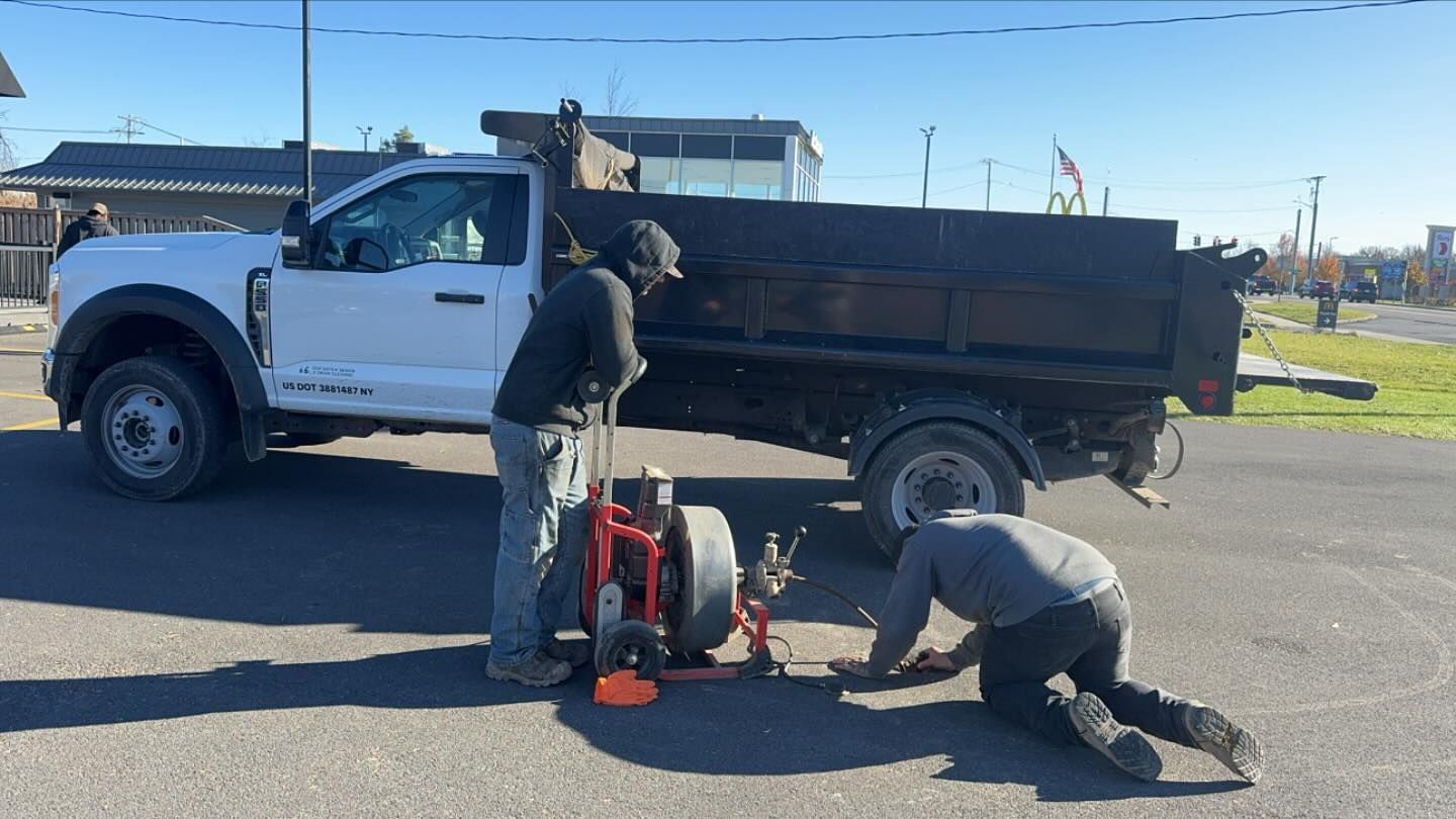 Two workers in a parking lot operate a piece of outdoor maintenance equipment next to a white dump truck.