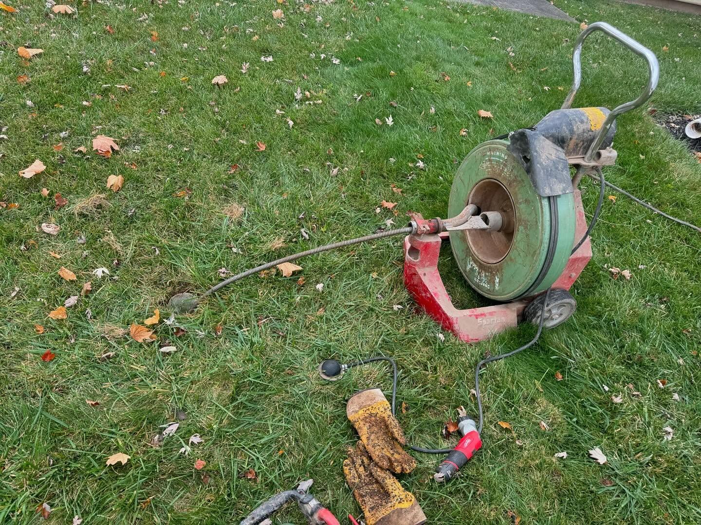 A drain cleaning machine with a coiled cable and a pair of work gloves sitting on a grassy lawn.