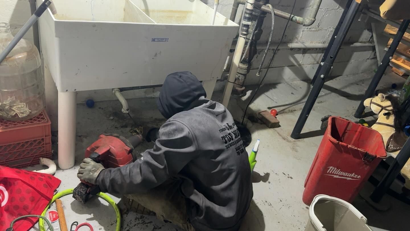 A person in a gray hooded sweatshirt uses a drain cleaning machine under a utility sink in a basement.