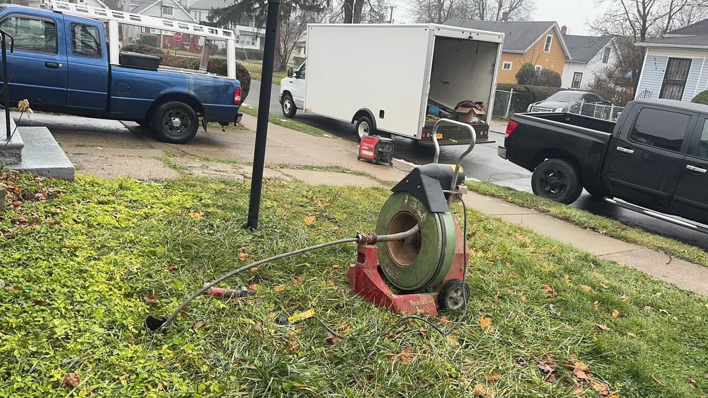 A drain cleaning machine sits on a residential lawn, with service trucks and a trailer parked on the street nearby.
