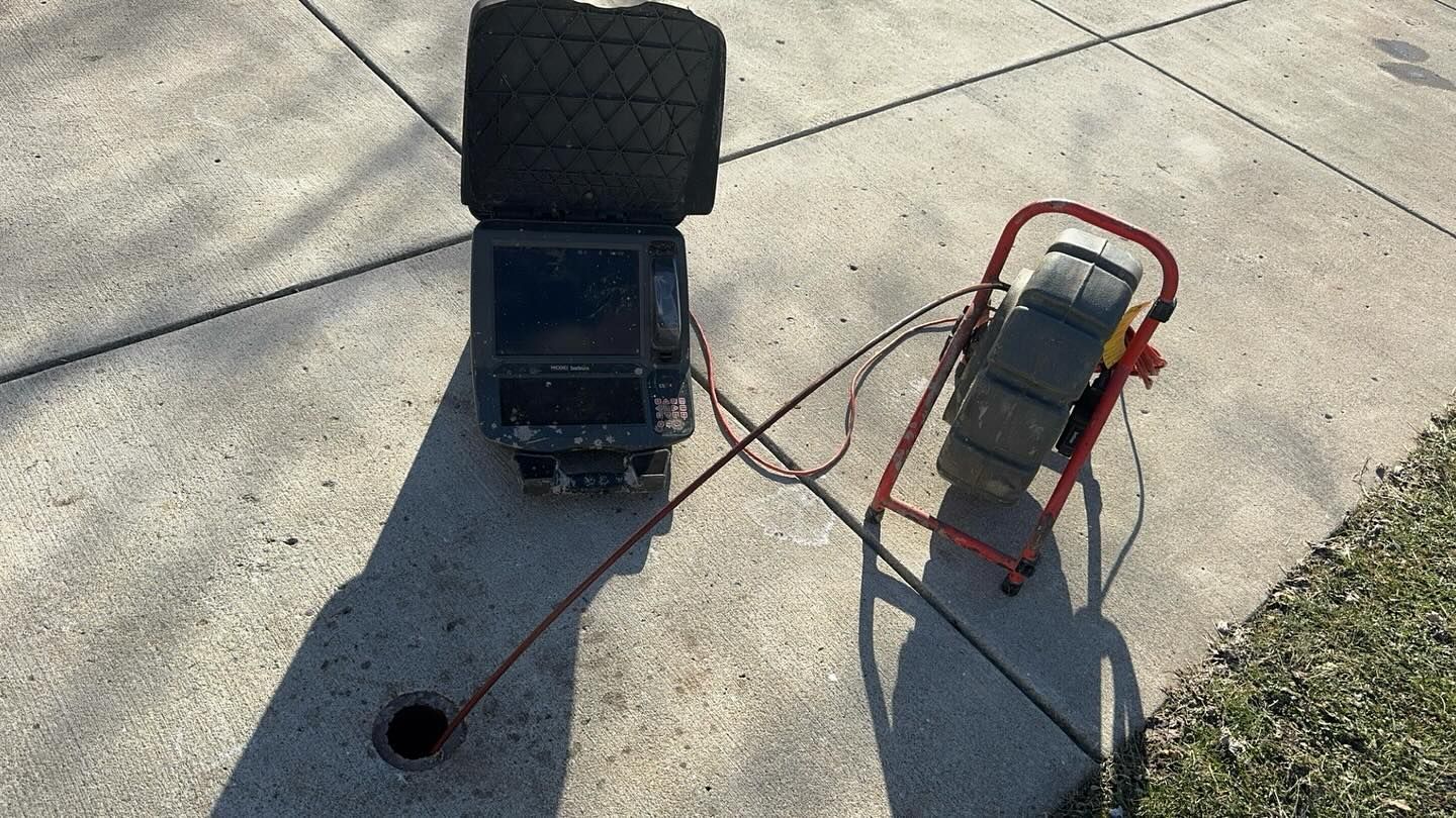 A sewer camera system with a monitor and a cable reel, connected to a circular drain opening in a concrete driveway.